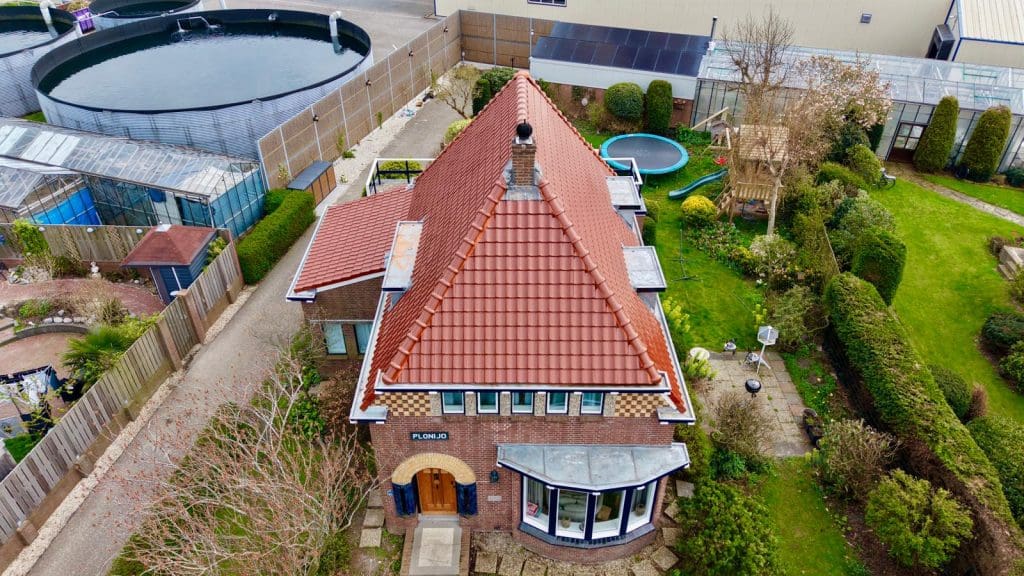 Bakstenen gebouw met een steil rood zadeldak met dakpannen, schoorsteen en bomen op de voorgrond.