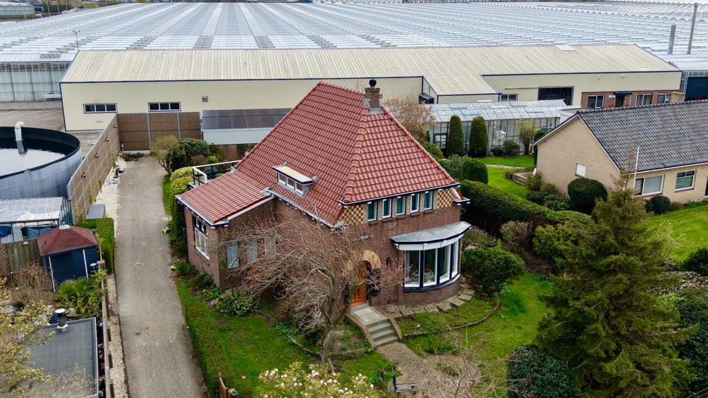 Luchtfoto van een woning met rood pannendak in schilddakvorm, omgeven door een groene tuin en bijgebouwen.