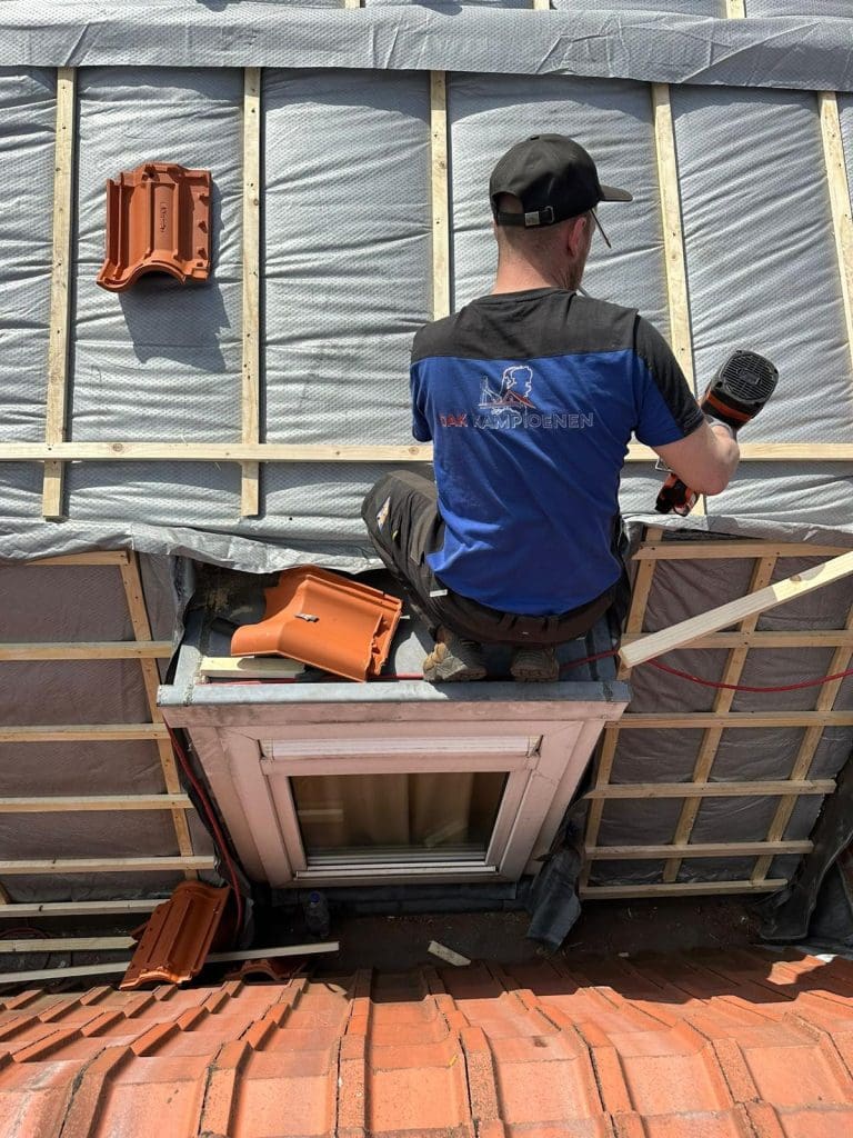 Persoon op een oud houten plat dak met dakdoorvoeren, schoorstenen en naastgelegen rood pannendak in een stedelijke woonomgeving.